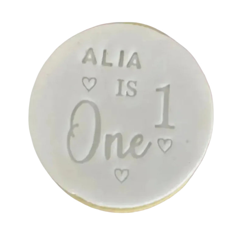 A round, white custom keepsake coin or medallion engraved with the text 'ALIA IS One 1' and small heart details, set against a solid pink background.
